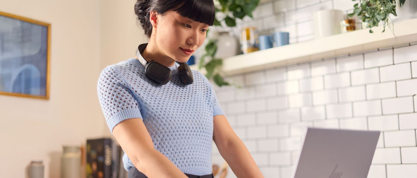 Woman with HP Poly headset around her neck working at a laptop in a kitchen (wide hero banner)